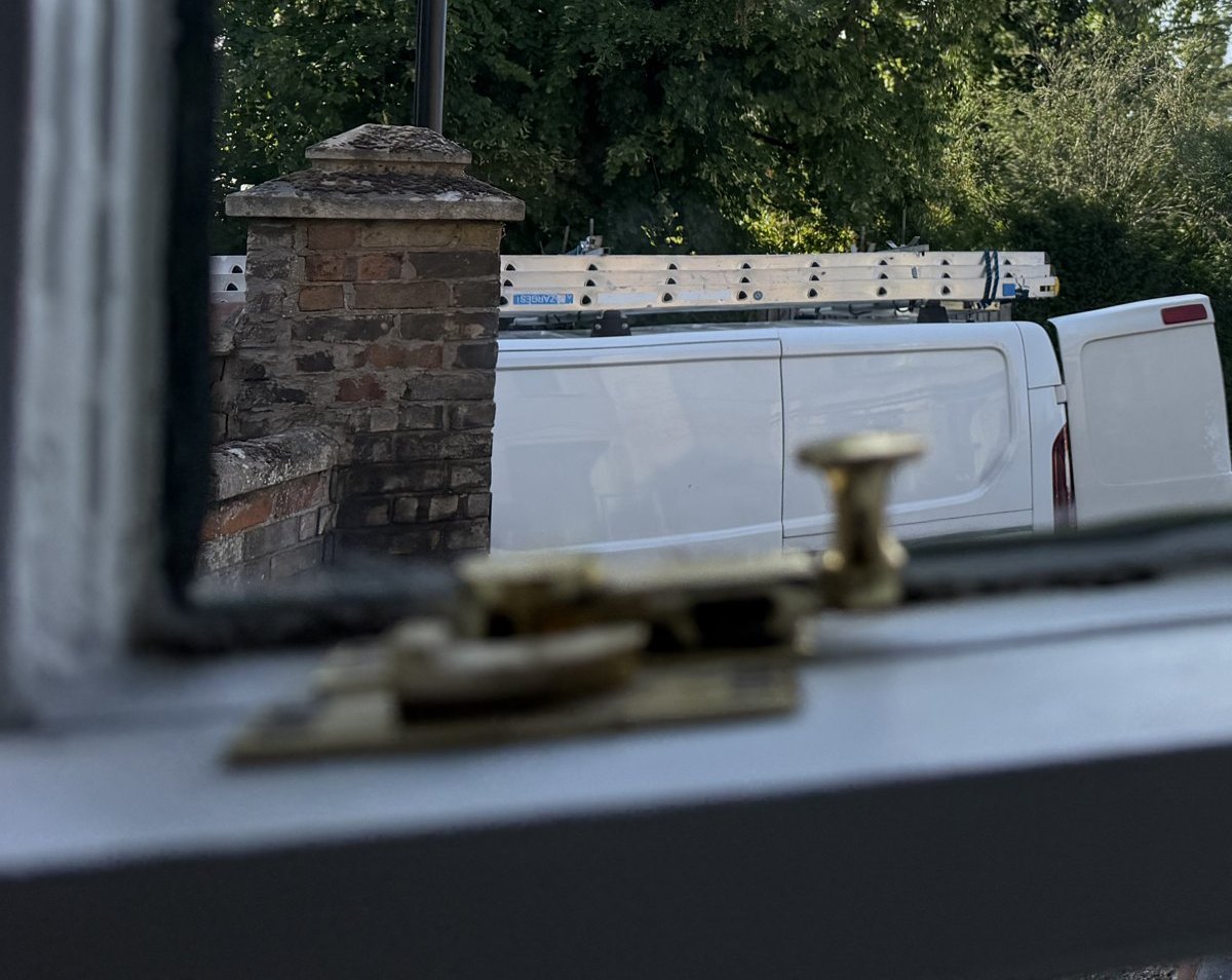 A brass sash fastener on the sill of a restored sash, seen from inside, a brick gate pier and a van beyond