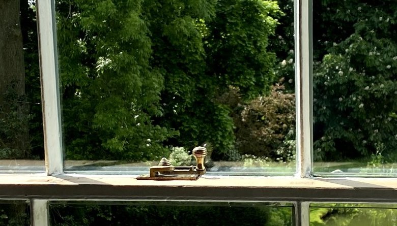 A brass sash fastener on the meeting rails of a cream-painted six-over-six sash, a garden beyond