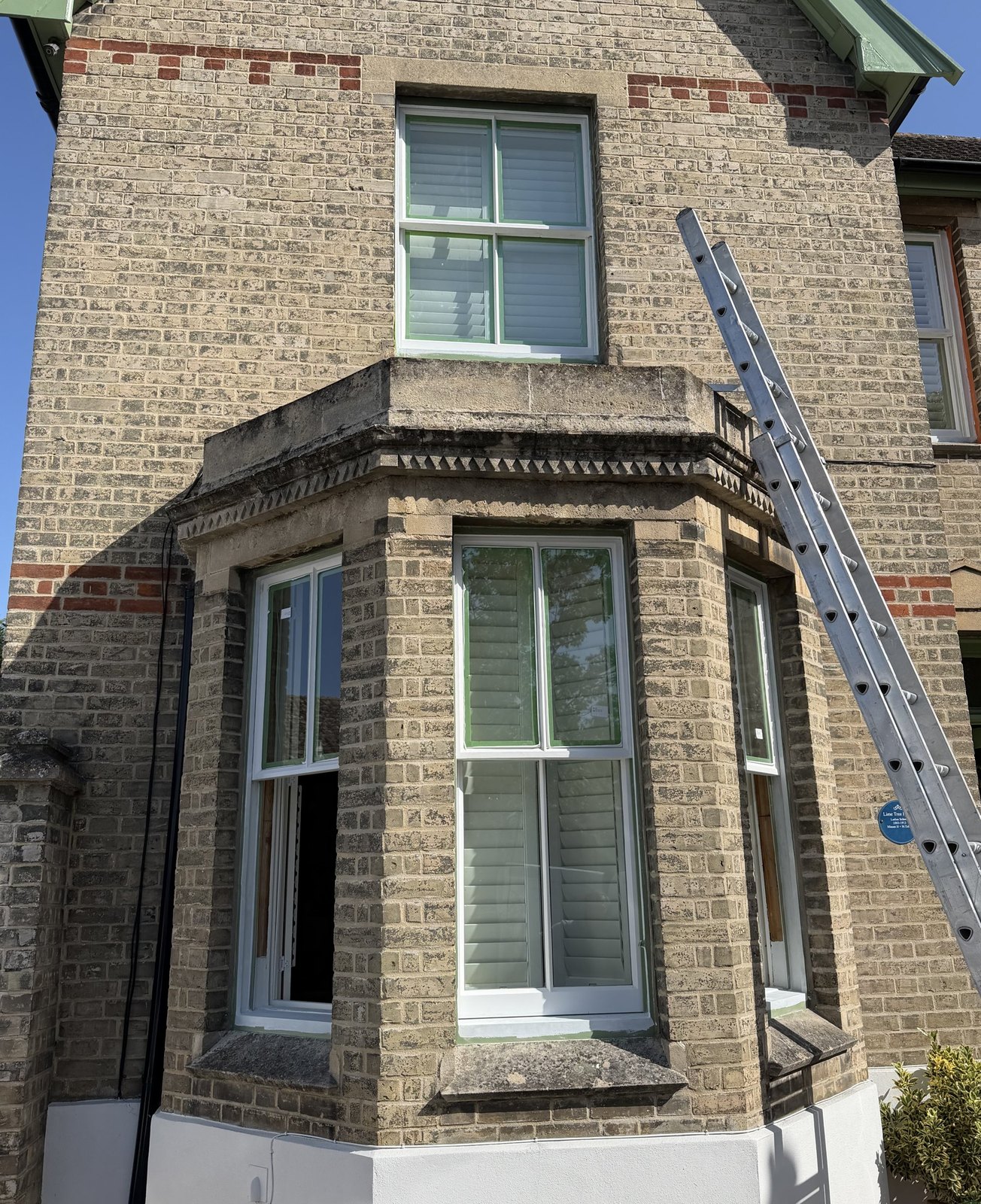 A gault-brick Victorian house with a canted bay of one-over-one sashes and a two-over-two sash above it, a ladder against the wall