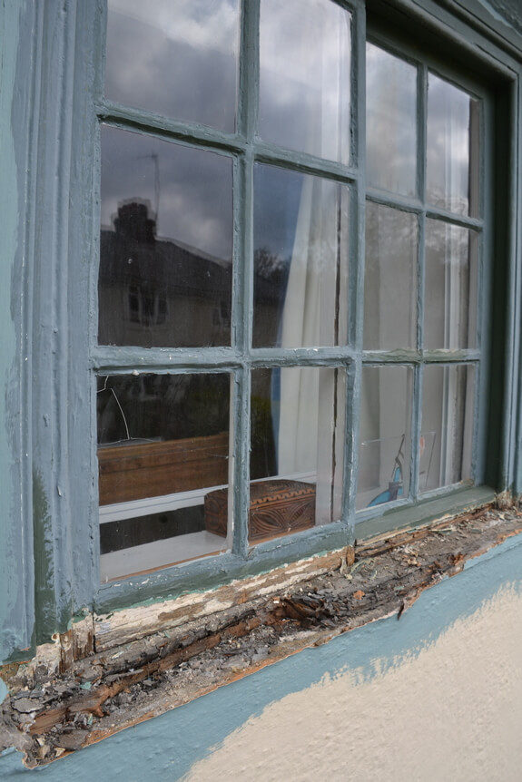 Badly rotten timber window sill with flaking blue-grey paint below a multi-pane window
