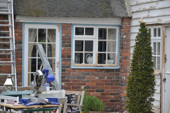 Mitre saw on a workbench outside a thatched brick cottage with a blue-framed casement window and glazed door