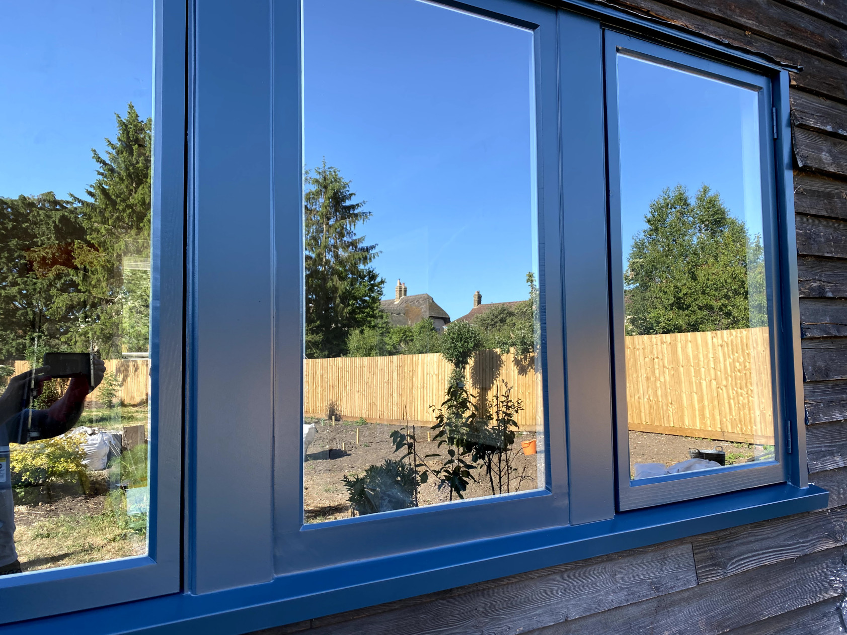 Close-up of a three-light timber casement window spray-painted blue in dark weatherboarding, reflecting the garden