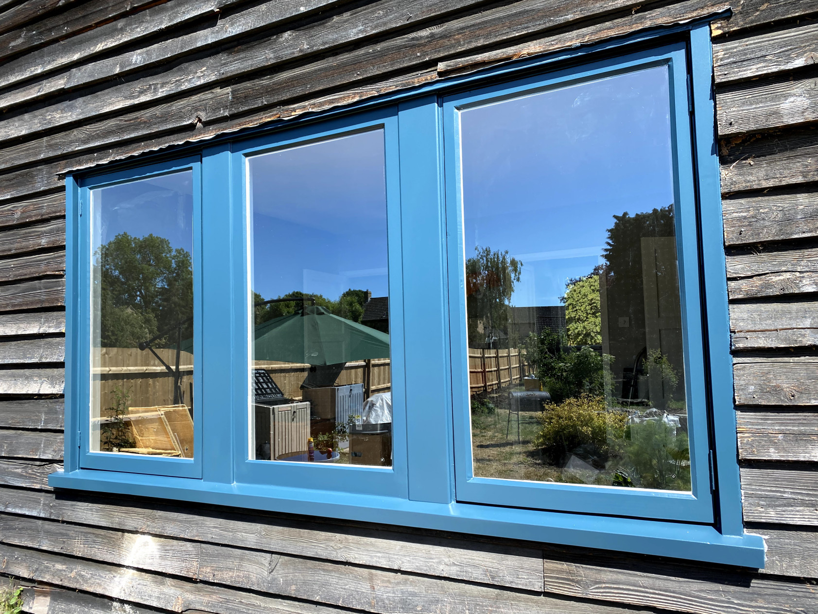 Three-light timber casement window spray-painted mid-blue, set in dark weathered timber cladding