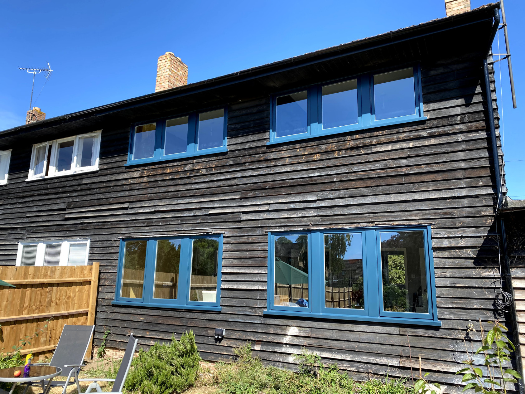 Two-storey house clad in dark weathered timber boarding with blue spray-painted timber casement windows