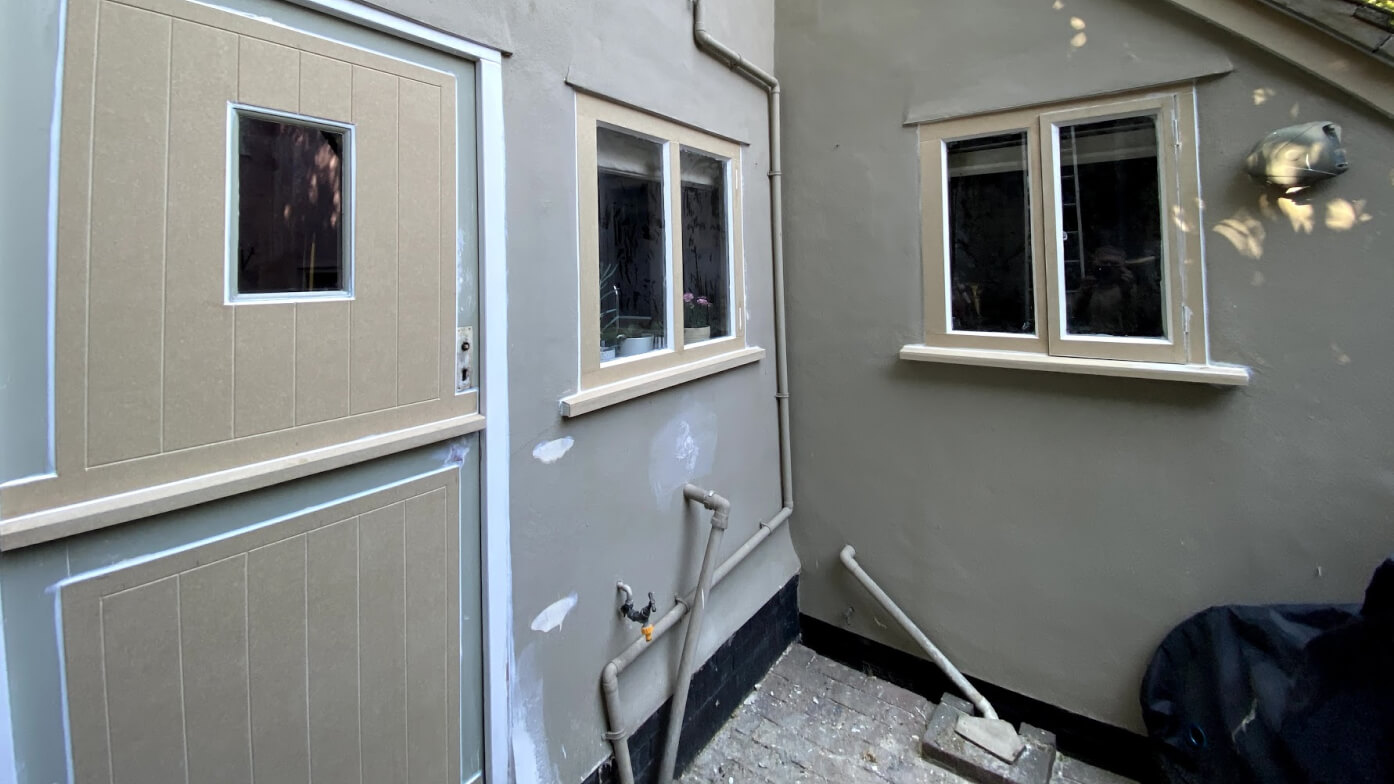 Boarded stable door and timber casement windows primed buff in a grey-rendered courtyard, walls dotted with filler
