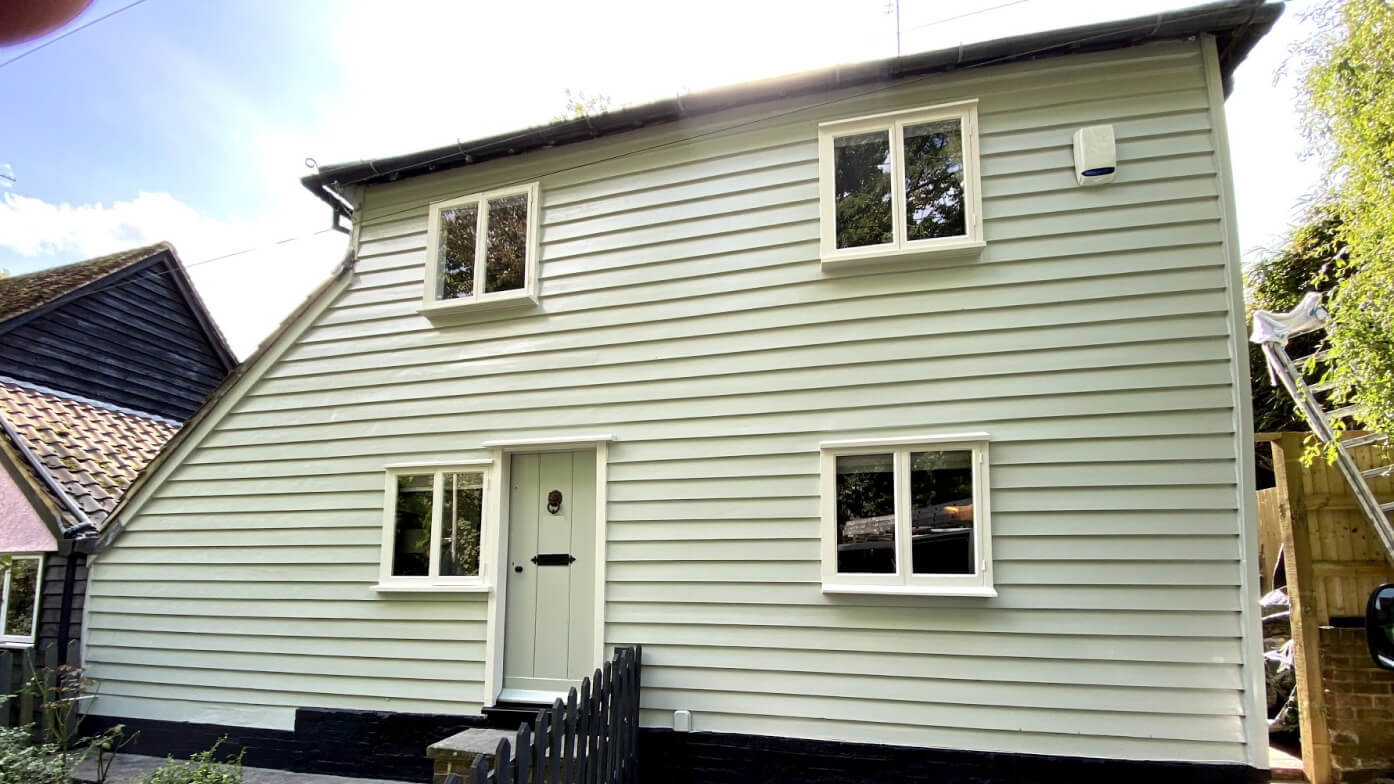 Weatherboarded house repainted pale sage green with white timber casement windows, a boarded front door and black plinth