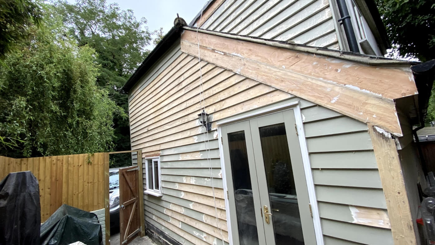 Weatherboarded house mid-restoration, with patchy grey-green cladding, a bare timber bargeboard and French doors