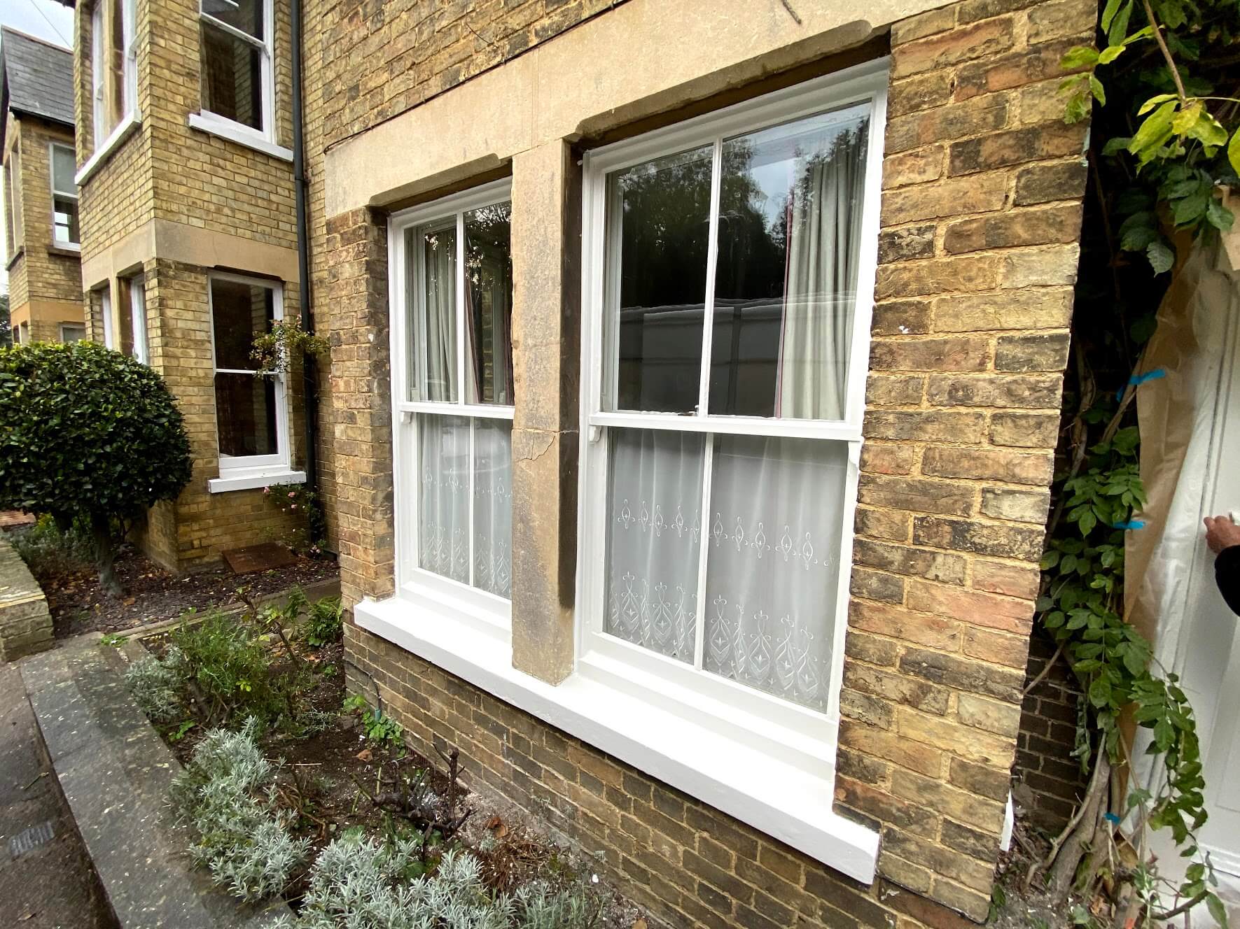 Two white-painted two-over-two sash windows beneath a stone lintel on the ground floor of a buff brick house