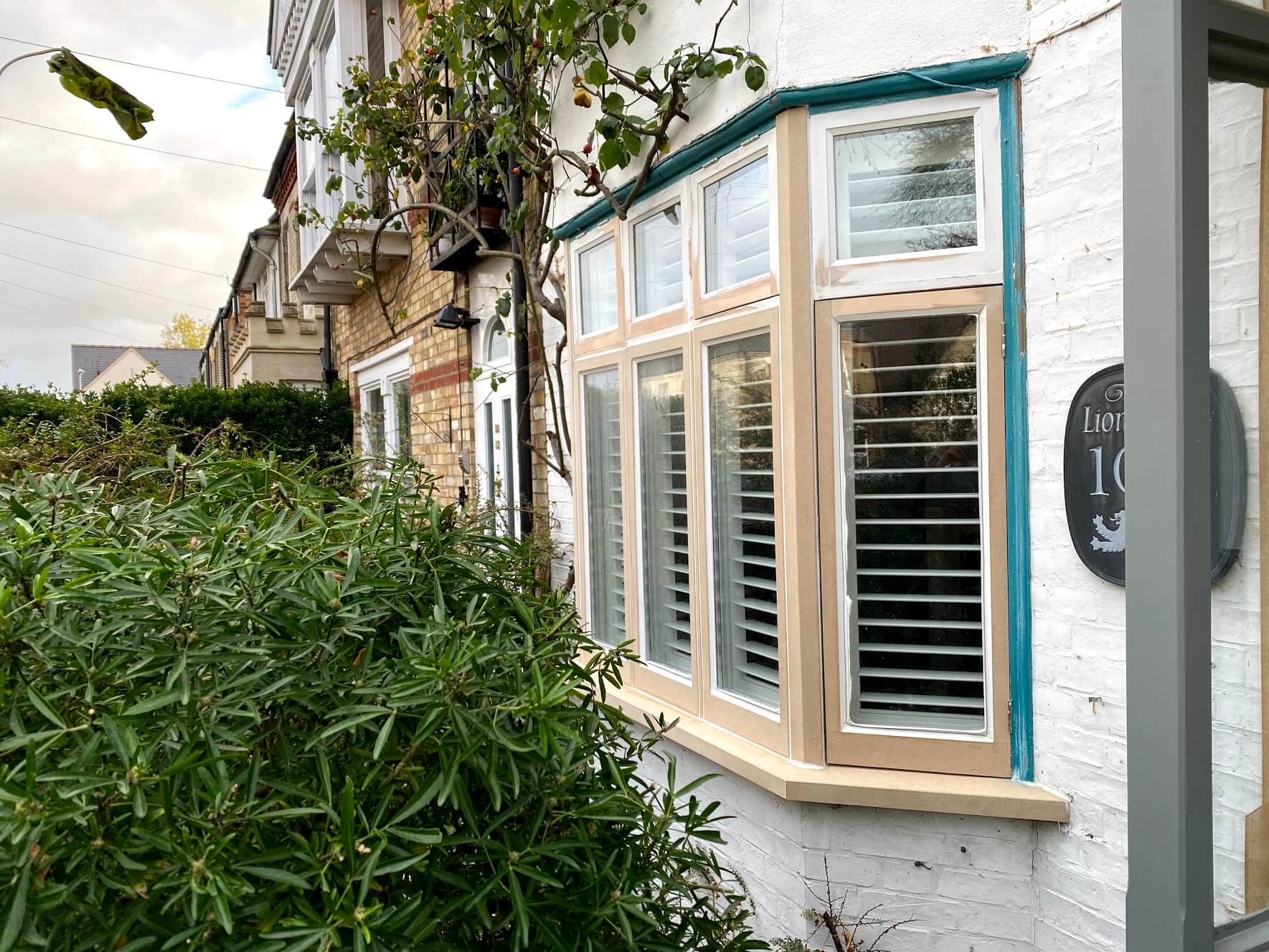 Timber casement bay window mid-restoration, frames coated pale beige inside a teal surround on white brickwork