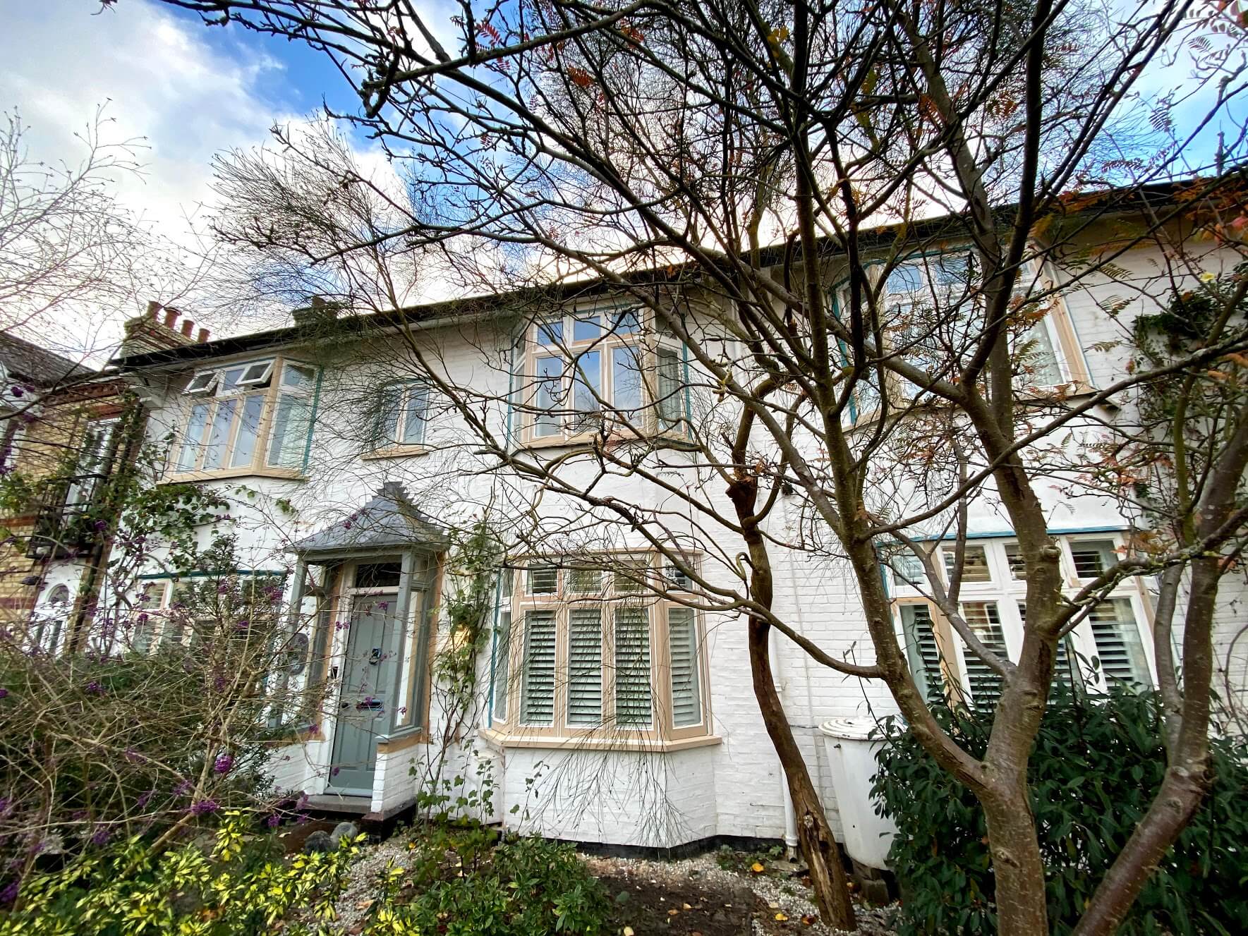 White-painted brick house with timber casement bay windows and a gabled entrance porch behind bare trees