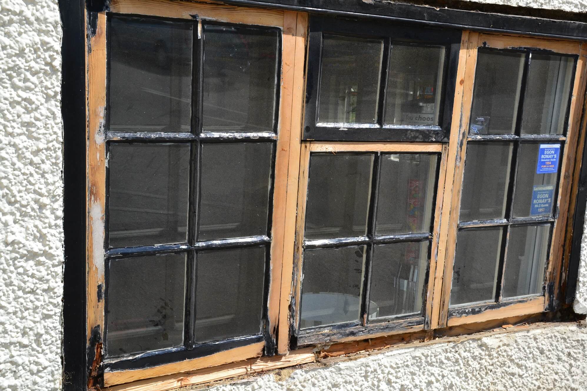 Small-paned casement window stripped back to bare timber, with the decayed sill exposed below, in a white-rendered wall