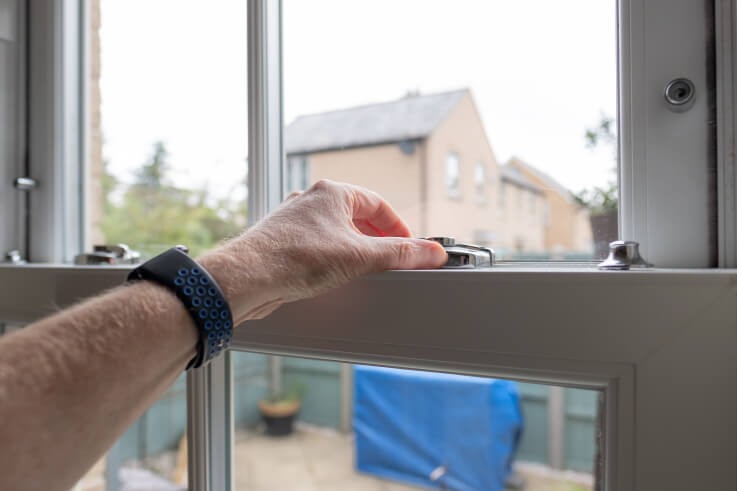 A hand lifting the lower sash of a white sash window by its lifts