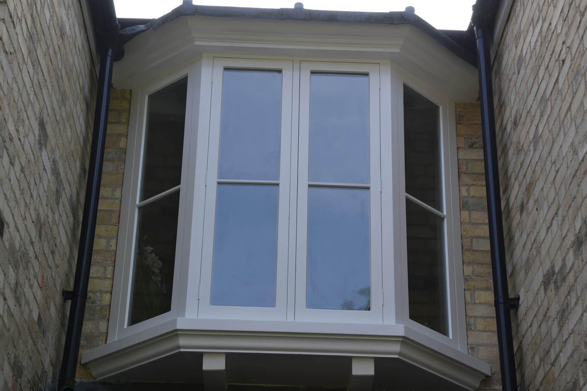 White-painted timber bay window with slim glazing bars, set between buff brick walls and black downpipes