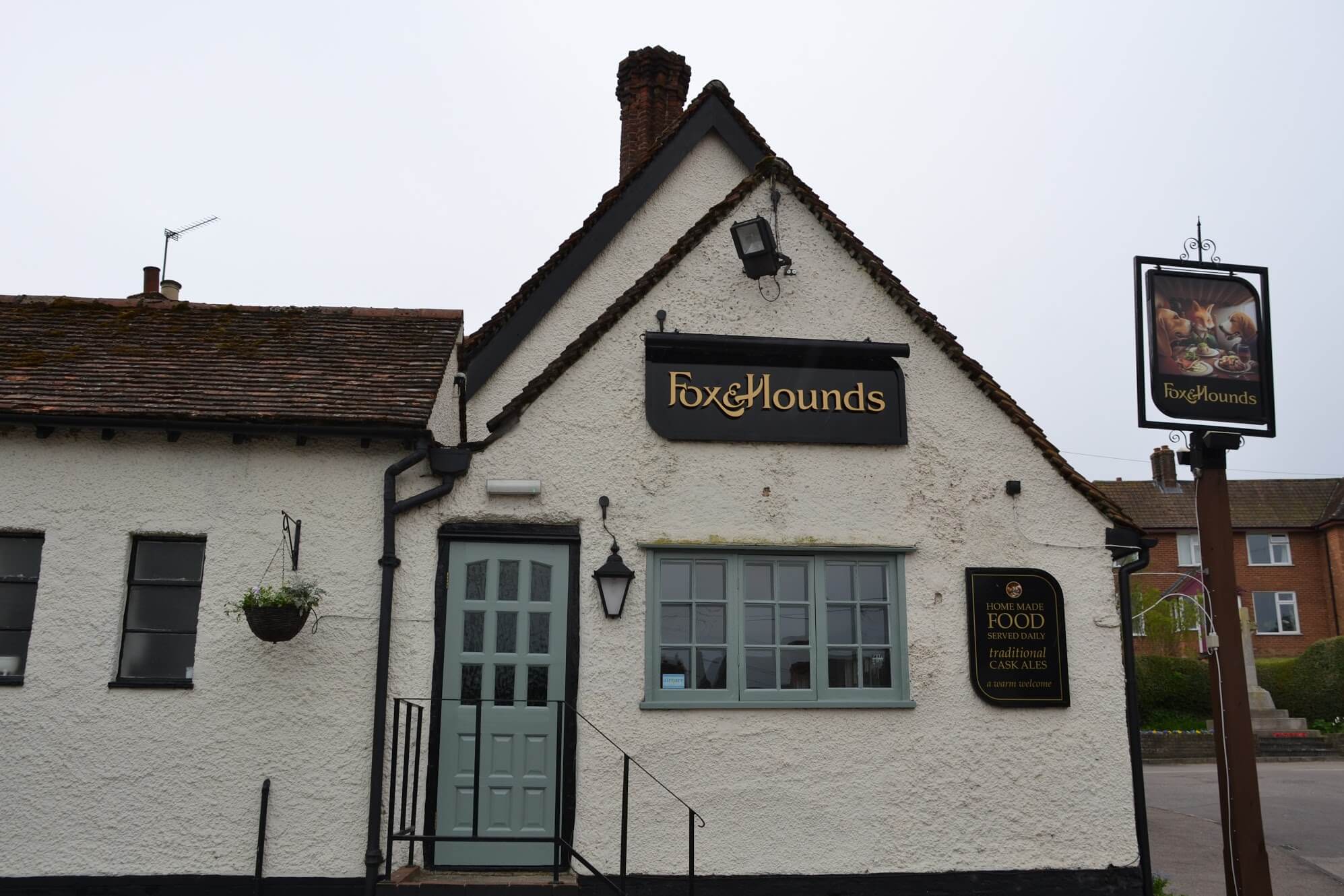 Front of the Fox & Hounds pub with its door and casement window repainted grey-green, white render and hanging sign