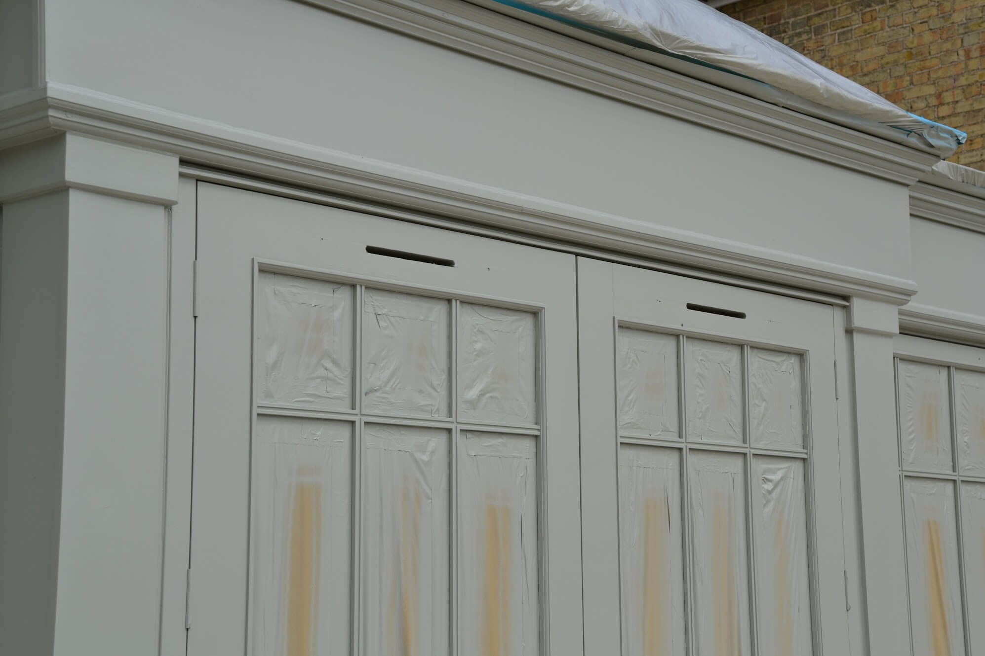 Timber conservatory doors and frame freshly spray-painted light grey, with the glass and roof masked in plastic