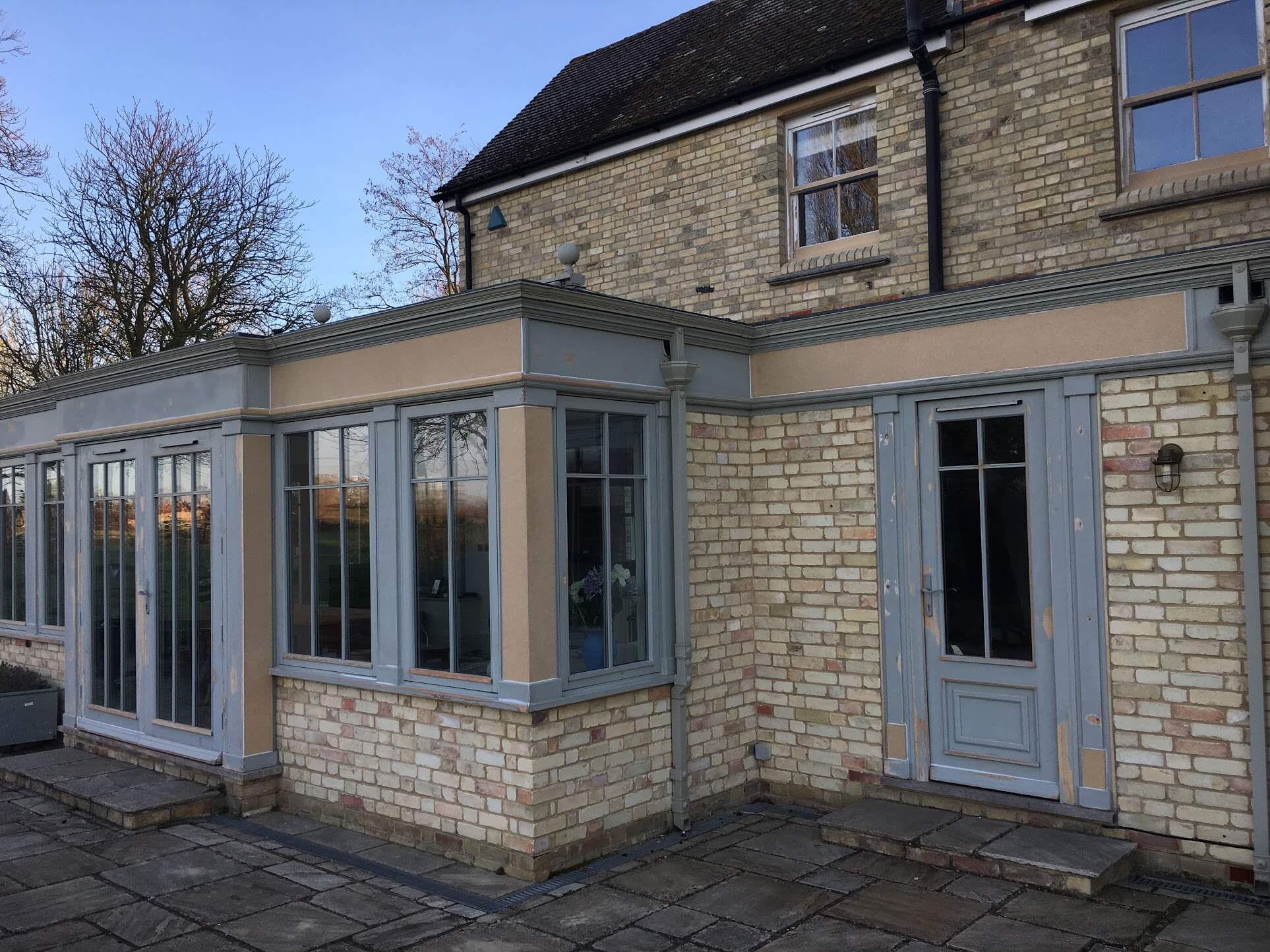 Grey-blue painted timber conservatory and garden door on a buff brick house, with beige primer patches on the fascia