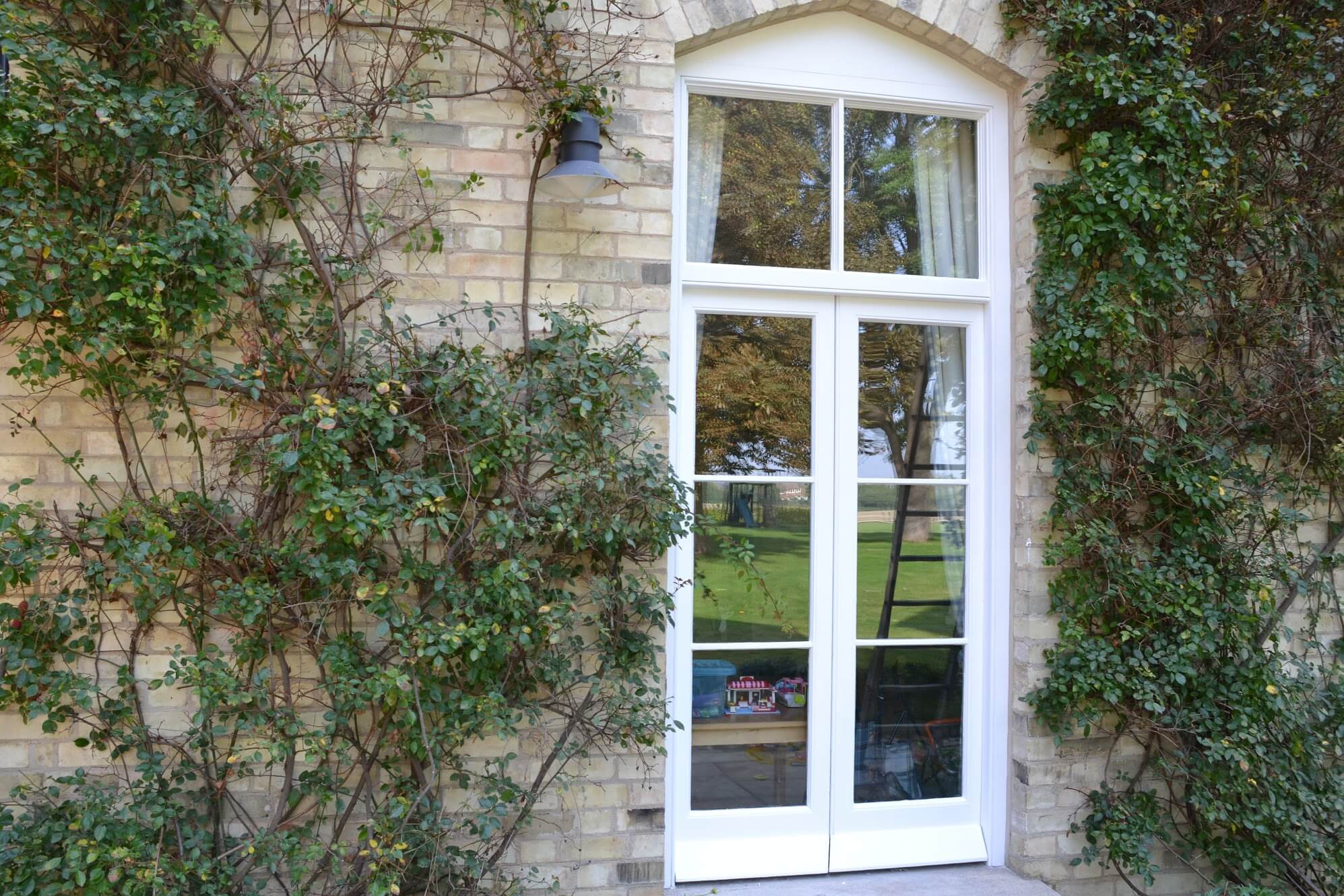 White-painted timber French doors beneath an arched fanlight in a buff brick wall framed by climbing plants