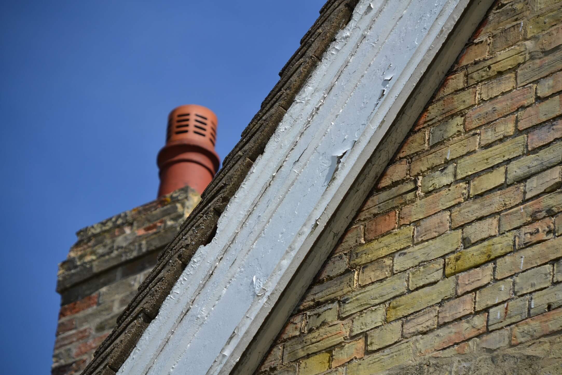 Flaking paint on a timber bargeboard along a brick gable, with a chimney pot behind