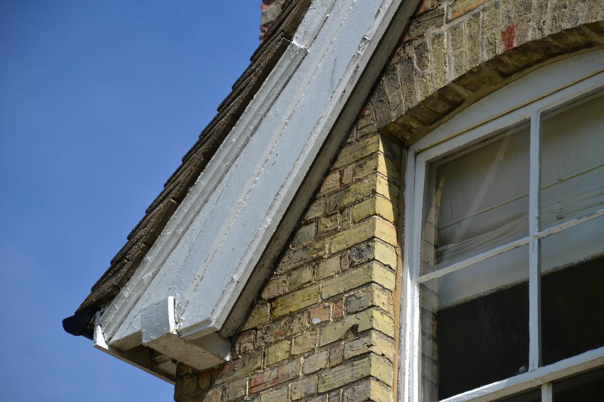 Weathered painted bargeboard at the foot of a brick gable beside an arched-head sash window
