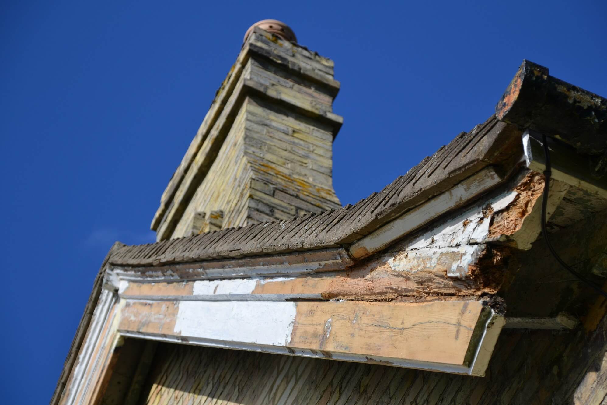 Rotten, crumbling end of a bargeboard at the eaves of a brick gable, with a chimney stack behind