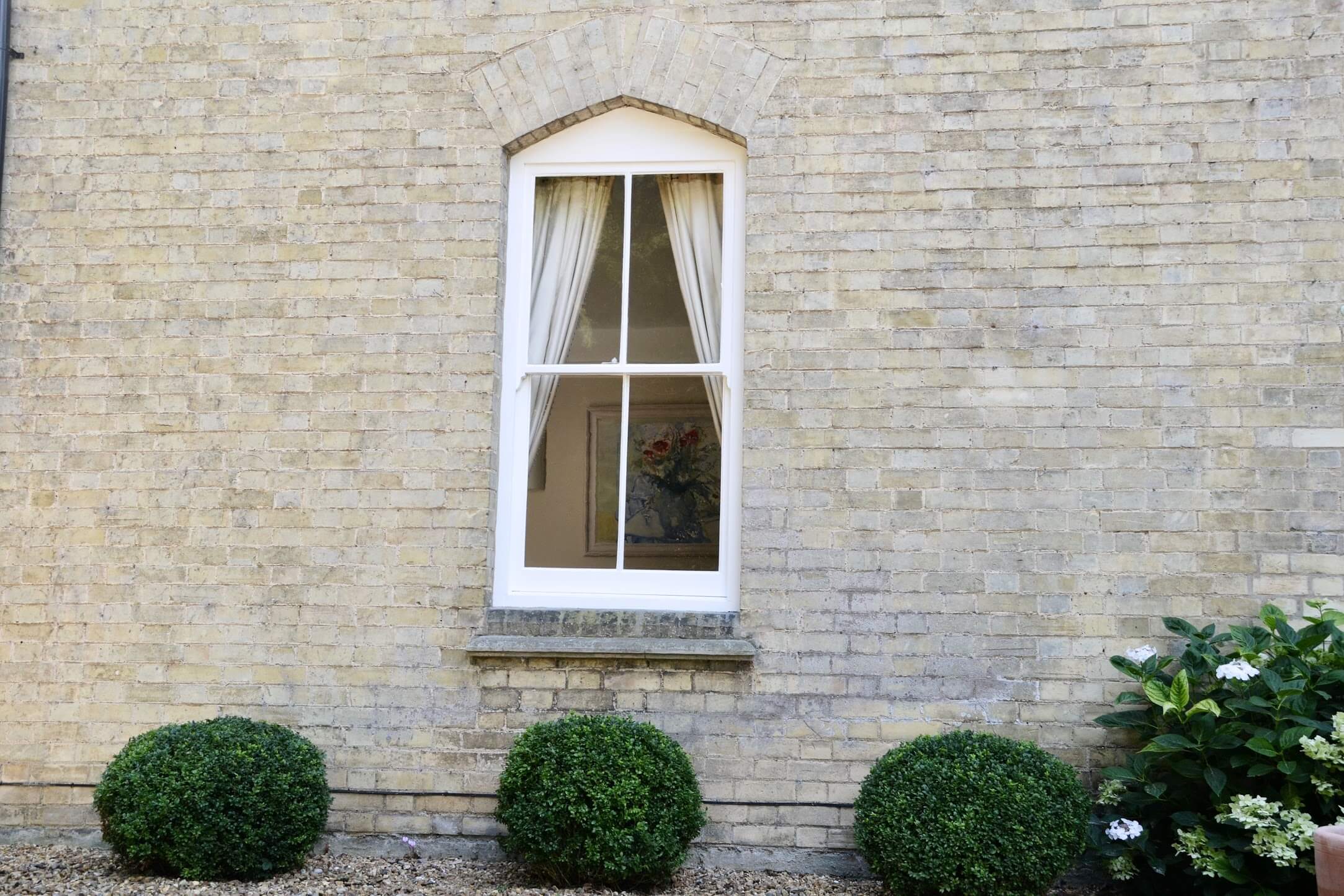 Victorian two-over-two sash window painted white under a brick arch in a buff brick wall, clipped box balls below