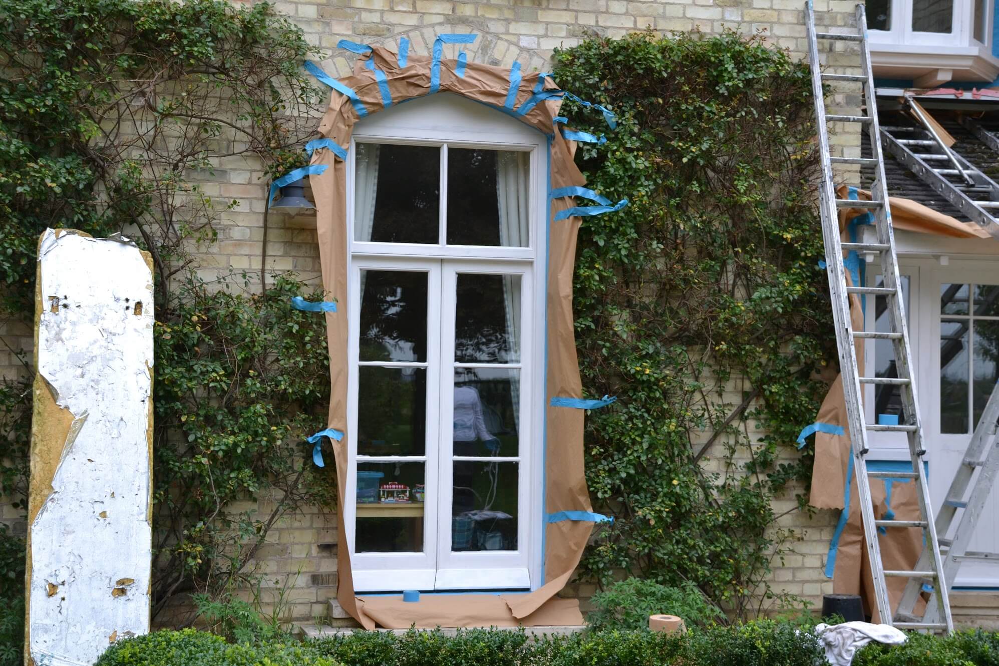 Tall timber casement window framed in brown masking paper and blue tape among climbing plants, ladders nearby