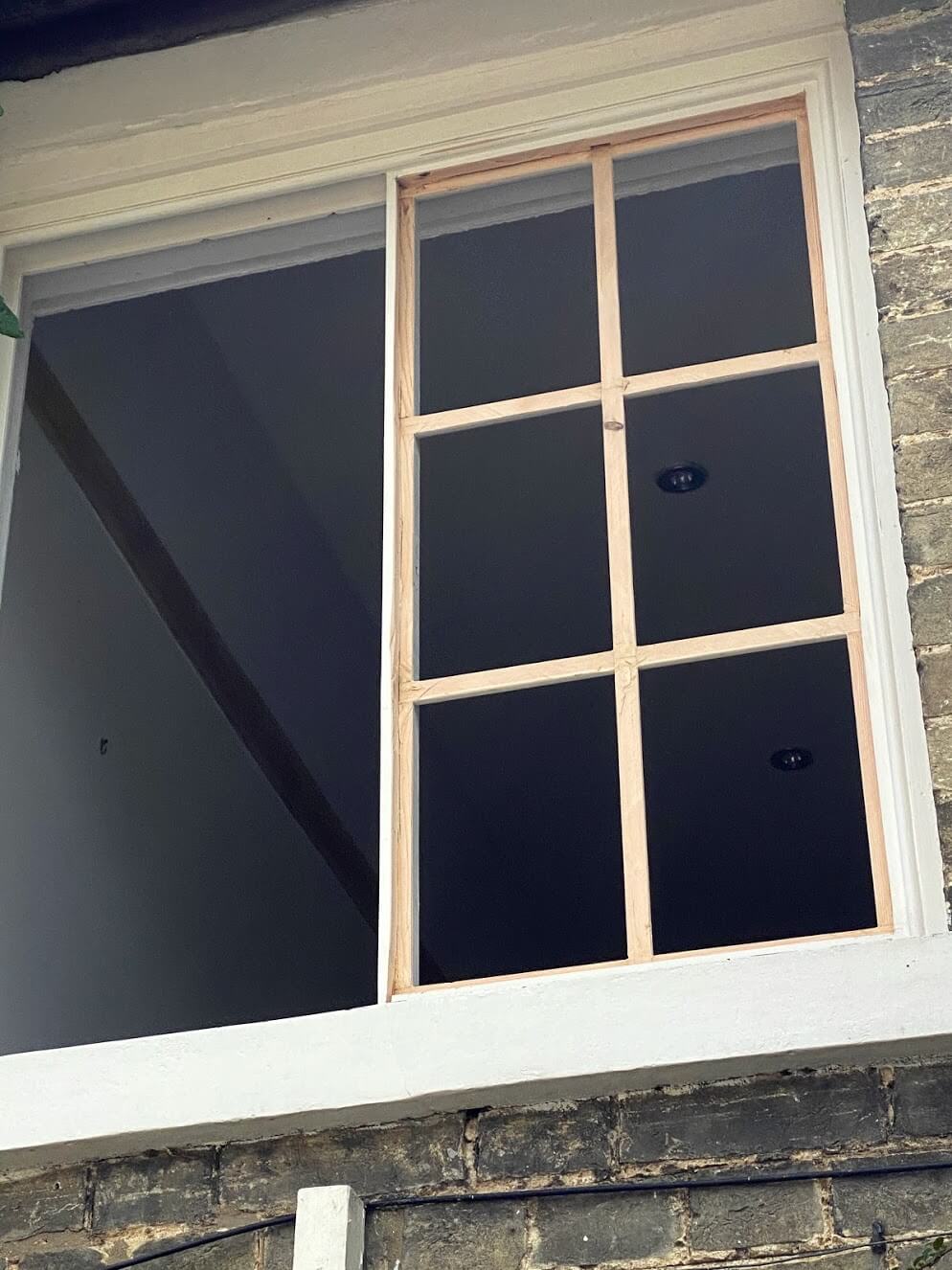 Bare timber six-pane Georgian sash in a white-painted frame during reglazing, beside an empty window opening