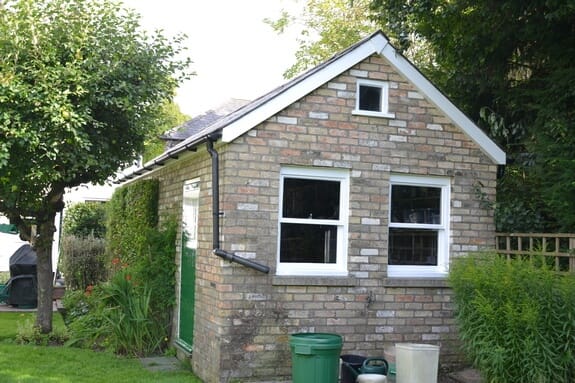 Buff brick garden outbuilding with two white sash windows, a green door and white bargeboards