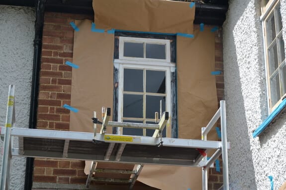Timber window framed in brown masking paper and blue tape behind a scaffold tower platform