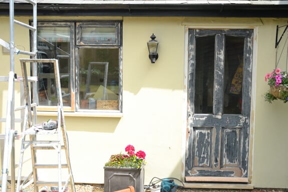 Cream-rendered cottage wall with a timber window and half-glazed door in flaking black paint, ladders set up alongside