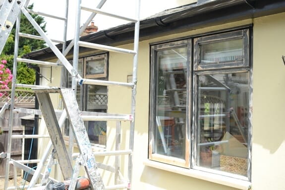 Scaffold tower beside timber windows partly sanded back to bare wood on a cream-rendered cottage wall