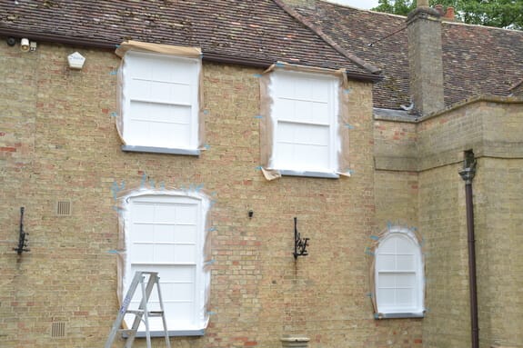 Pale brick house with its windows masked off in protective sheeting for painting, a stepladder below