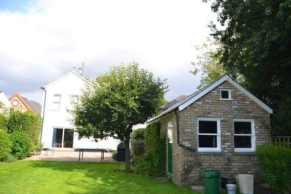Garden lawn with the white-painted house behind a tree and a buff brick outbuilding with white windows