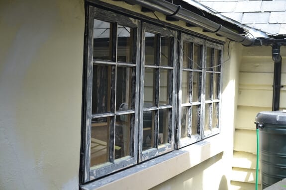 Row of four small-paned timber casement windows with worn, flaking black paint under a slate roof edge