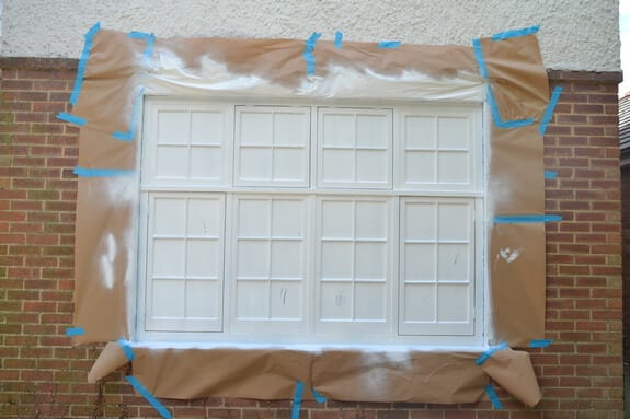Multi-pane casement window in a brick wall, sprayed white and surrounded by brown masking paper and blue tape