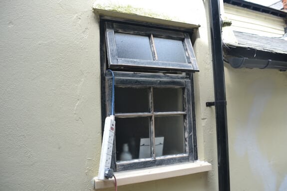 Small timber window with a frosted top light and worn black paint in a cream-rendered wall beside a downpipe