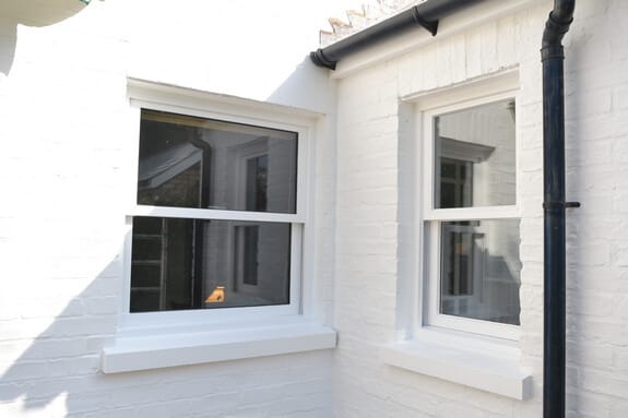 Two white one-over-one sash windows in white-painted brickwork beside a black downpipe