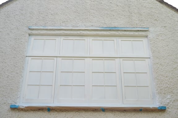 Wide multi-pane casement window sprayed white in a roughcast gable, glazing masked and blue tape on the sill