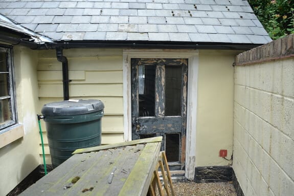 Half-glazed timber back door with flaking black paint under a slate roof, with a water butt and workbench in front