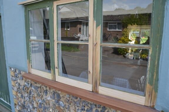Timber window partly stripped to bare wood, with a new unpainted sill fitted above a flint wall