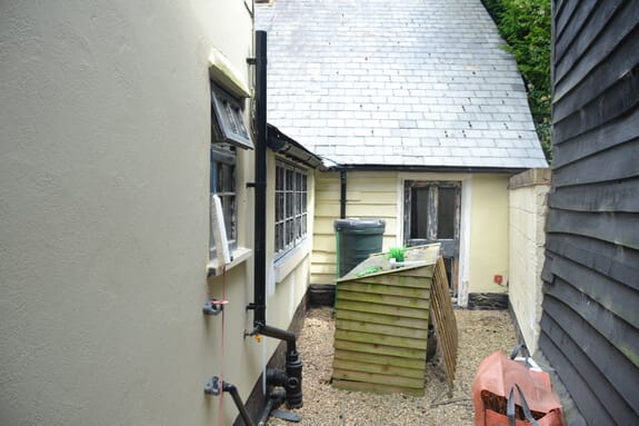 Narrow gravel passage between a cream-rendered cottage and black weatherboarding, with timber windows along the wall