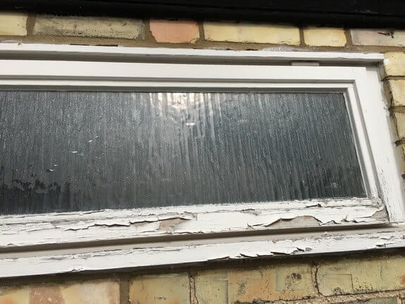 Small obscure-glazed timber window in yellow brick with badly peeling white paint on the bottom rail and sill