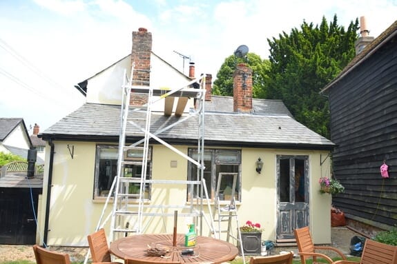 Cream-rendered cottage with slate roof and brick chimneys, a scaffold tower set up in front of its timber windows