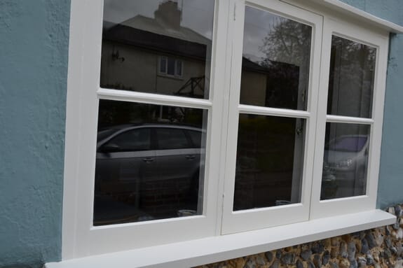 Timber window with slim glazing bars repainted off-white, above a flint wall
