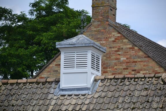 White louvred cupola with a lead-covered roof and weathervane on a tiled roof by a brick chimney
