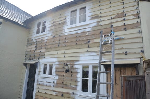 Two-storey weatherboarded cottage wall sanded back to bare wood in patches, with a ladder and white casement windows