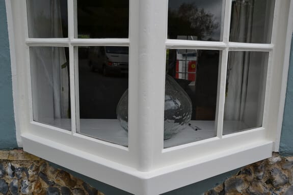Corner of a small timber bay window repainted white above flint walling, a glass vase inside