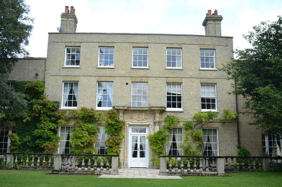 Garden front of a pale brick house with white sash windows on three floors and a carved stone doorcase