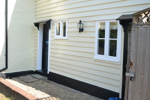 Cream weatherboarding with white casement windows, a black front door with canopy, a lantern and a black plinth