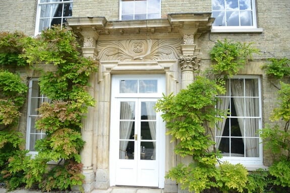 White glazed garden doors in a carved stone surround, flanked by sash windows and climbing plants